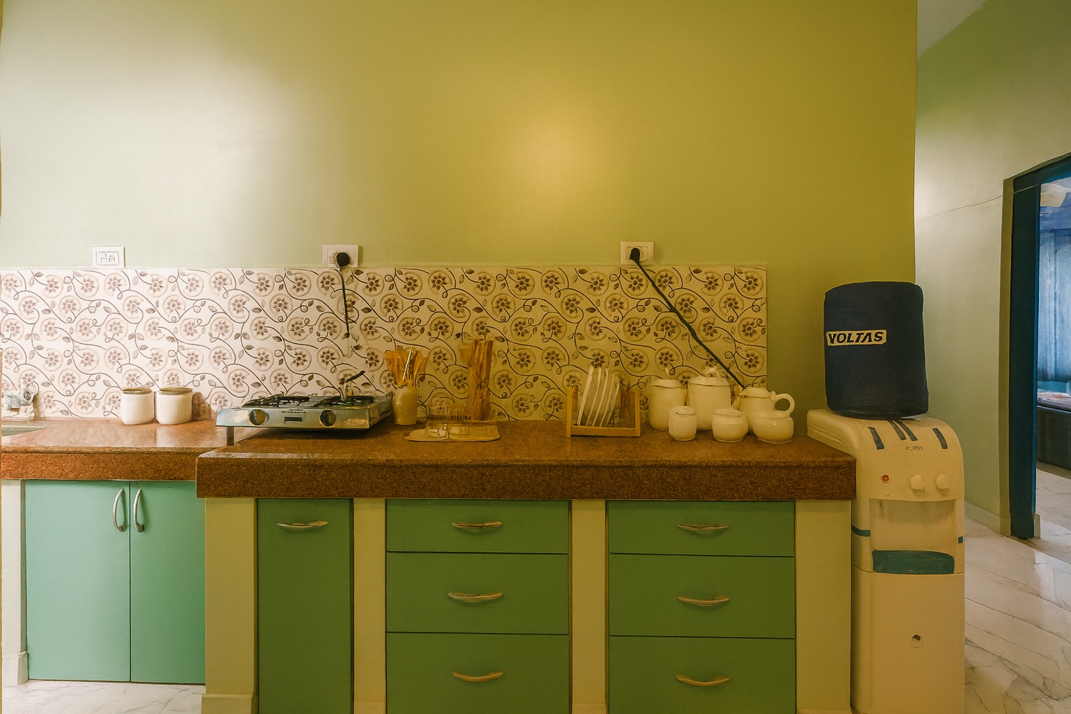Boutique, Varanasi: kitchen counter with a two-burner gas stove, jars and jugs against a floral-tiled wall, green cabinets and a water dispenser