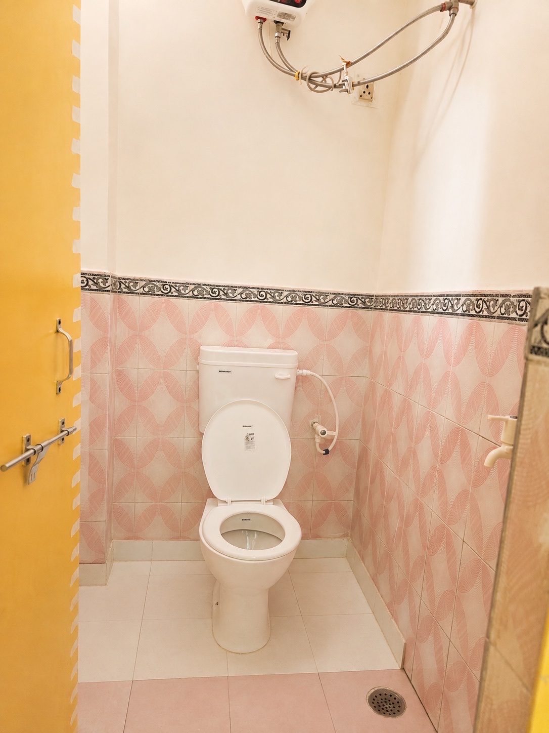Gardenia, Varanasi: bathroom with a western toilet and health faucet, pink patterned tiling under a scrolled border, water heater above