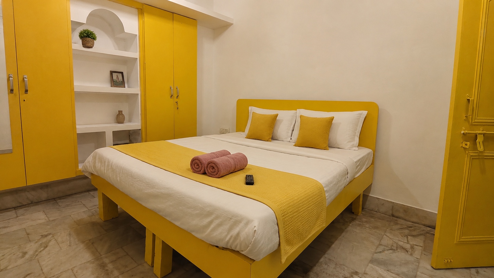Gardenia, Varanasi: double bed with a yellow headboard and bedspread, rolled towels laid out, mustard wardrobes either side of a white arched niche, marble floor