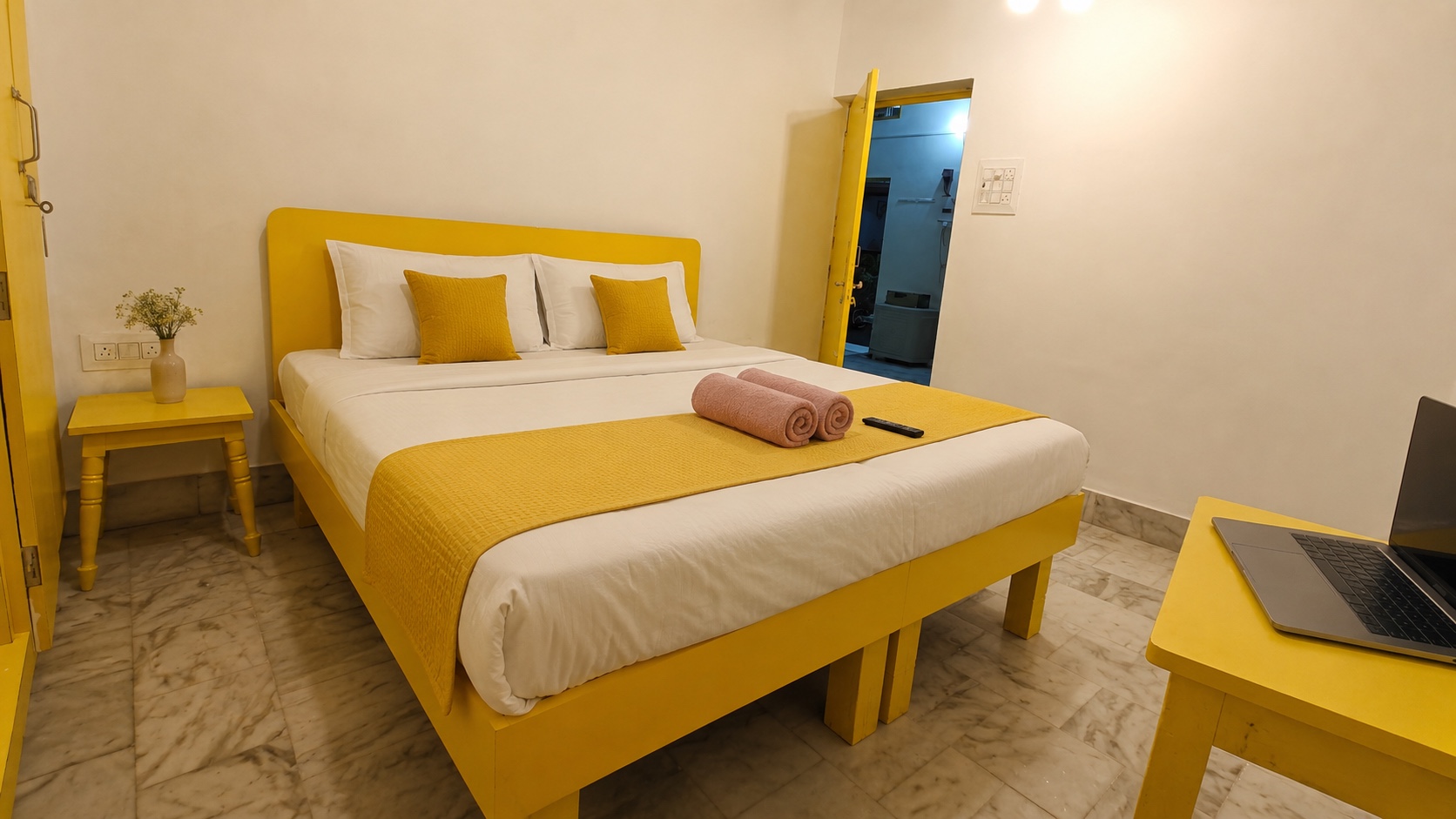 Gardenia, Varanasi: yellow-framed double bed with a turned bedside table and a laptop on a yellow desk, door open to the corridor