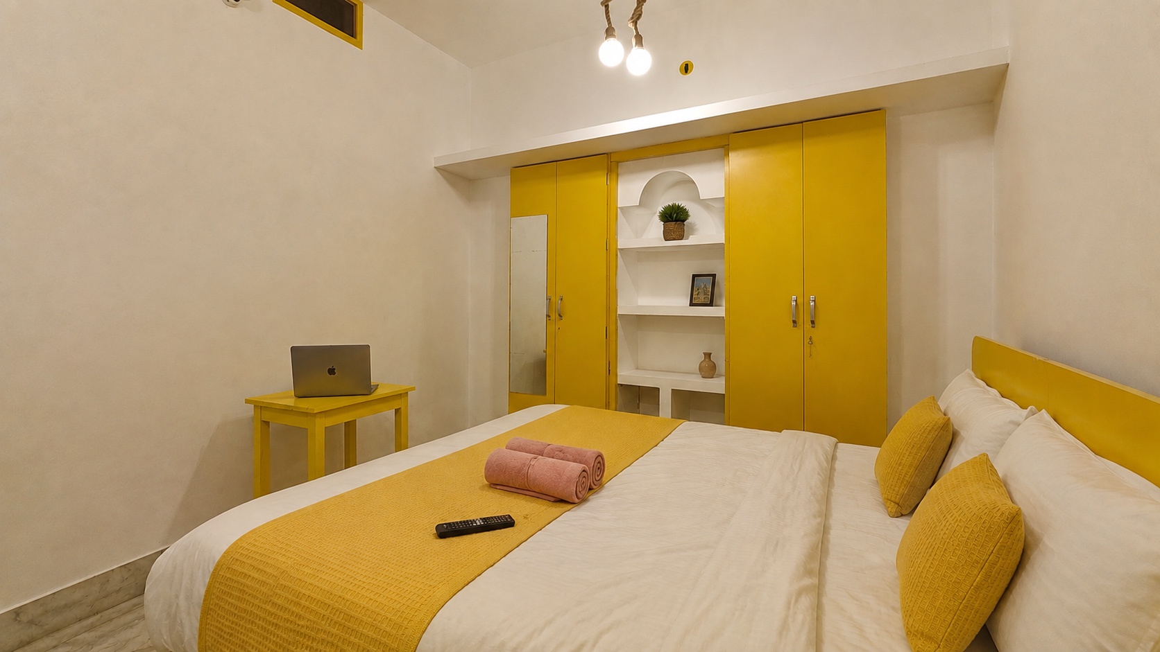 Gardenia, Varanasi: double bed facing a wall of mustard wardrobes with an open shelf of ornaments, laptop on a yellow writing table