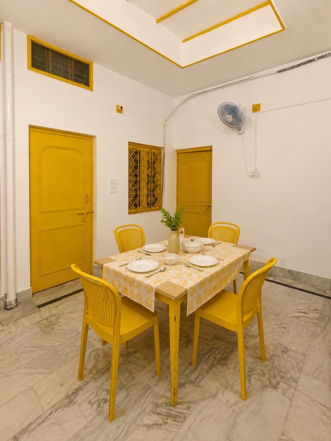Gardenia, Varanasi: dining table laid for four with yellow chairs and a printed cloth, mustard doors and a wall fan, marble floor under a coffered ceiling