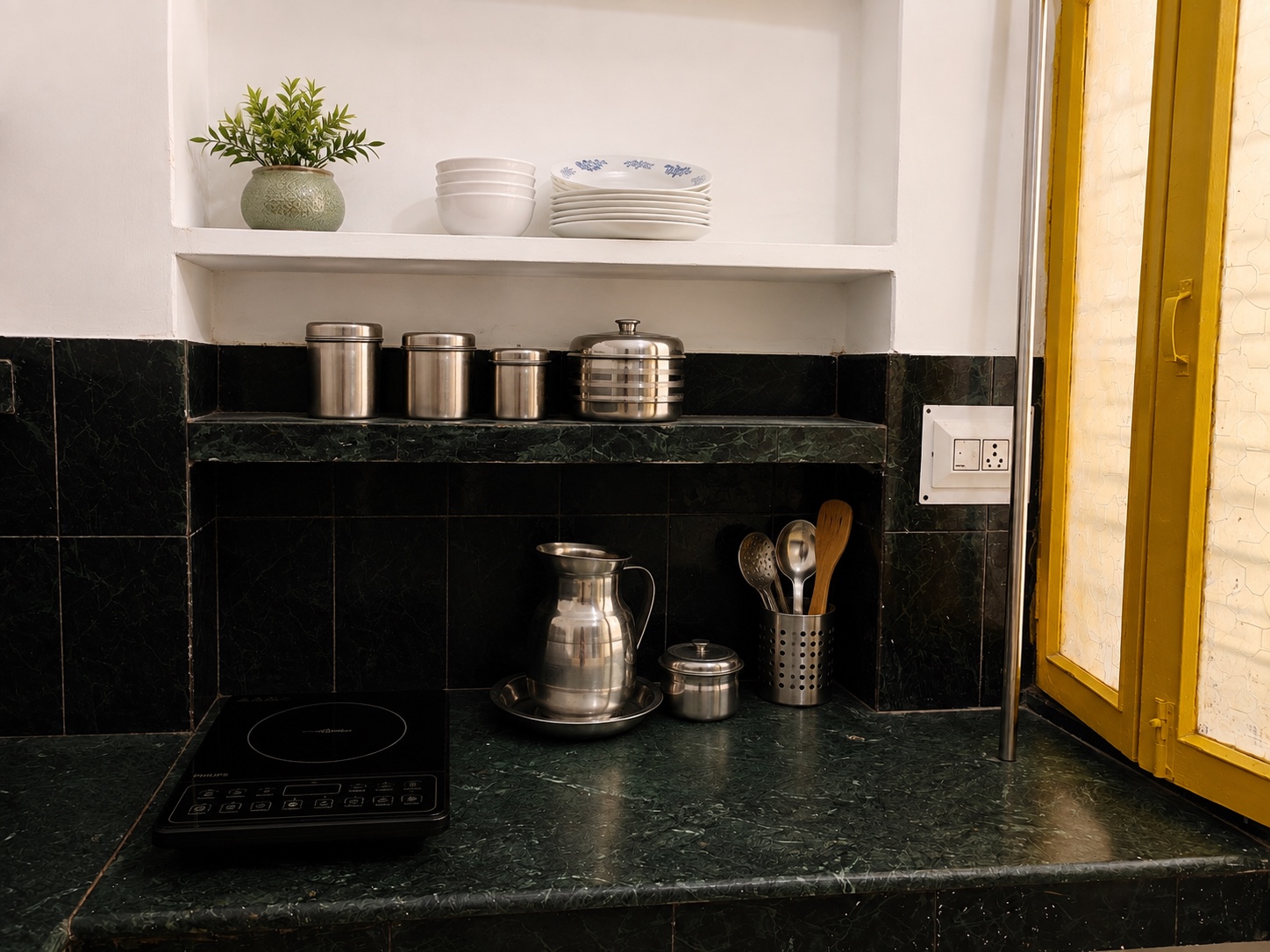 Gardenia, Varanasi: induction hob on the green marble counter with a steel water jug, spice tins and wooden utensils, beside a yellow glazed door