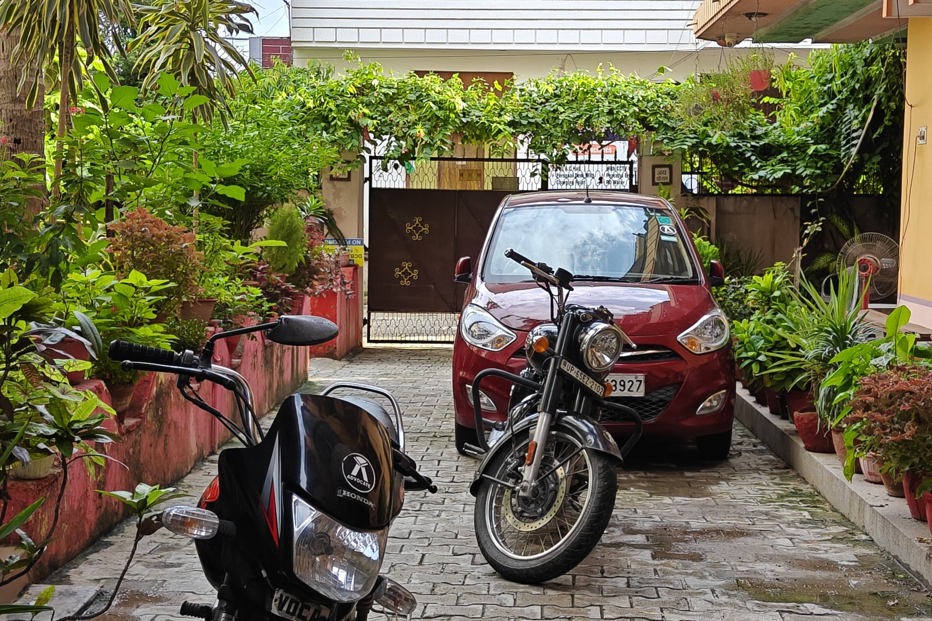 Gardenia, Varanasi: paved parking between potted plants, a car and two motorcycles inside the gate, creeper along the boundary