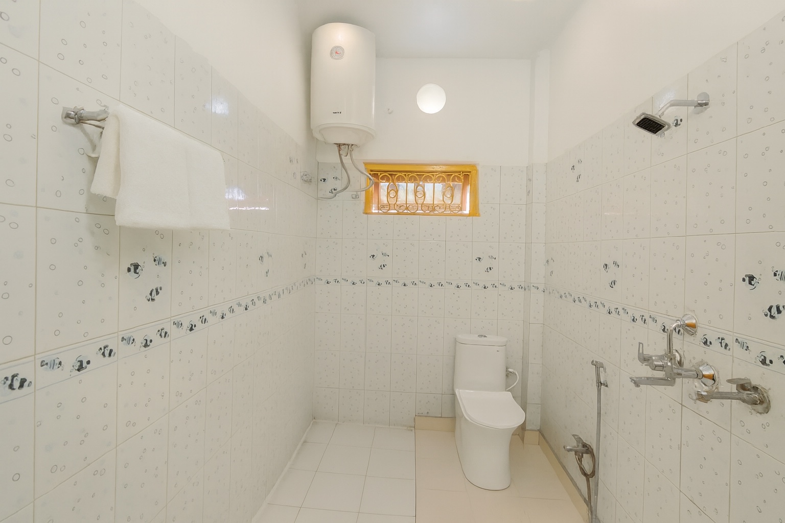 Minimilist, Varanasi: tiled bathroom with an overhead water heater, wall-mounted shower, western toilet with a health faucet and a white towel on the rail