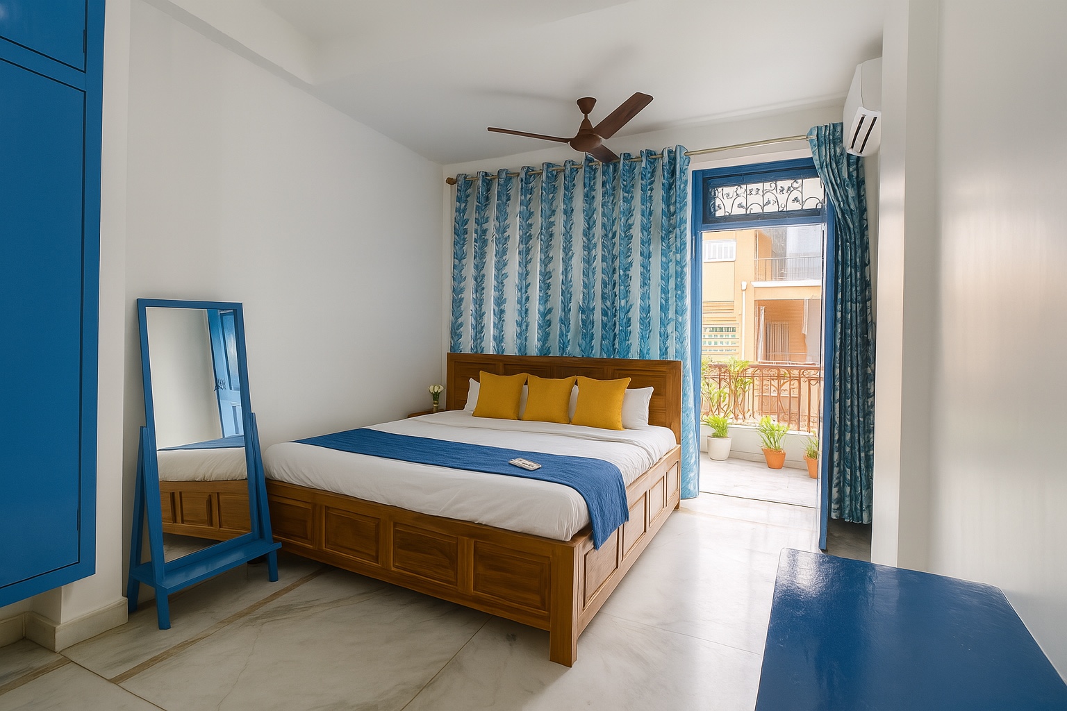 Santorini, Varanasi: white room with mustard cushions on a teak bed, a blue-framed standing mirror and doors open to a balcony of potted plants